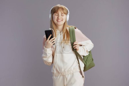 Foto de Feliz adolescente moderna en chándal beige con mochila escuchando la música con auriculares y utilizando aplicaciones de teléfonos inteligentes contra gris. - Imagen libre de derechos