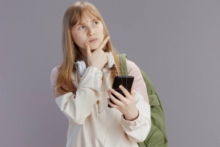 Photo for Pensive trendy school girl in beige tracksuit with backpack and headphones using smartphone app isolated on grey background. - Royalty Free Image