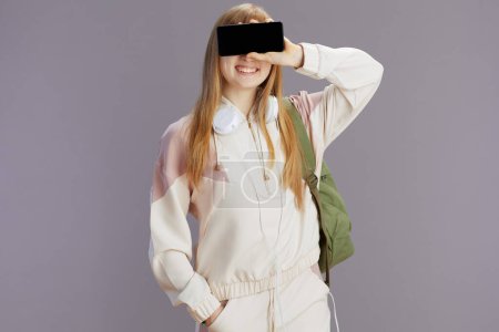 Photo for Smiling trendy pupil in beige tracksuit with backpack and headphones showing smartphone blank screen isolated on grey background. - Royalty Free Image