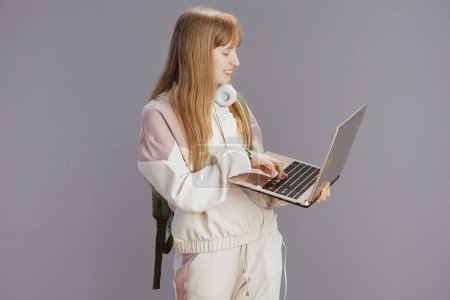 Téléchargez les photos : Fille à la mode souriante en survêtement beige avec écouteurs à l'aide d'un ordinateur portable isolé sur gris. - en image libre de droit