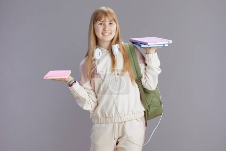 Photo for Portrait of happy modern young woman in beige tracksuit with backpack, workbooks and headphones isolated on grey background. - Royalty Free Image