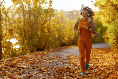Photo for Hello autumn. Full length portrait of modern female in fitness clothes in the park listening to the music with headphones and jogging. - Royalty Free Image
