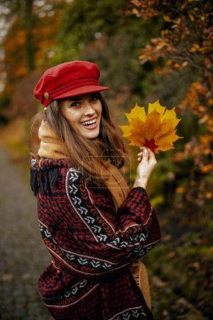 Téléchargez les photos : Bonjour l'automne. Portrait d'une élégante femme d'âge moyen souriante au chapeau rouge avec des feuilles d'automne et une écharpe marchant dans le parc de la ville. - en image libre de droit