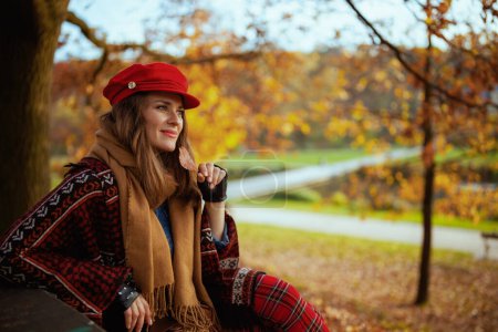 Photo for Hello autumn. pensive modern woman in red hat with autumn leaf and scarf sitting on bench in the city park. - Royalty Free Image