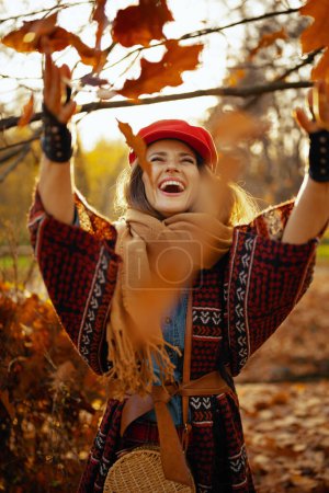 Photo for Hello autumn. smiling young woman in red hat with scarf and gloves throwing autumn leafs in the city park. - Royalty Free Image