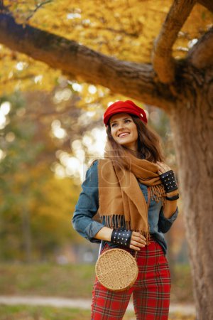 Téléchargez les photos : Bonjour l'automne. femme souriante moderne de 40 ans en jeans chemise et chapeau rouge avec écharpe, gants et sac près de l'arbre dans le parc de la ville. - en image libre de droit