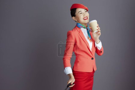 Foto de Sonriente elegante aire azafata asiático mujer en rojo falda, chaqueta y sombrero uniforme con café taza y rueda bolsa aislado en gris. - Imagen libre de derechos