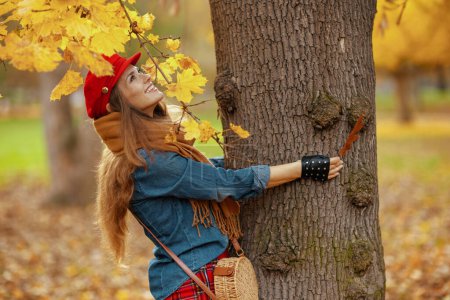 Foto de Hola otoño. mujer elegante sonriente en camisa de jeans y sombrero rojo con hoja de otoño, bufanda, guantes y bolso cerca del árbol en el parque de la ciudad. - Imagen libre de derechos