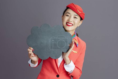 Photo for Smiling elegant asian female flight attendant in red skirt, jacket and hat uniform showing blank cloud shape board isolated on gray. - Royalty Free Image