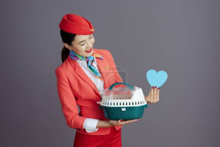 Téléchargez les photos : Souriant moderne hôtesse asiatique femme en jupe rouge, veste et chapeau uniforme avec animal de compagnie porteur montrant coeur isolé sur gris. - en image libre de droit