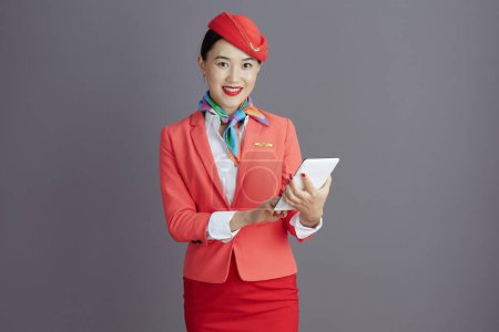 Téléchargez les photos : Souriant élégant hôtesse de l'air asiatique femme en jupe rouge, veste et chapeau uniforme avec tablette PC sur fond gris. - en image libre de droit