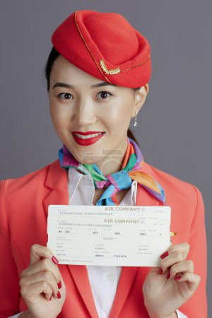 Téléchargez les photos : Heureux moderne asiatique femme hôtesse de l'air en jupe rouge, veste et chapeau uniforme avec billets d'avion isolé sur gris. - en image libre de droit