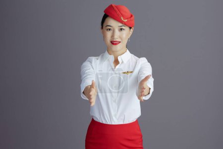 Photo for Stylish asian female stewardess in red skirt and hat uniform showing where the exits are against gray background. - Royalty Free Image
