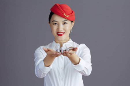 Photo for Smiling modern asian female stewardess in red skirt and hat uniform with a little airplane isolated on gray. - Royalty Free Image