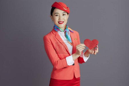 Téléchargez les photos : Heureux élégant hôtesse de l'air asiatique femme en jupe rouge, veste et chapeau uniforme avec du papier rouge coeur isolé sur fond gris. - en image libre de droit
