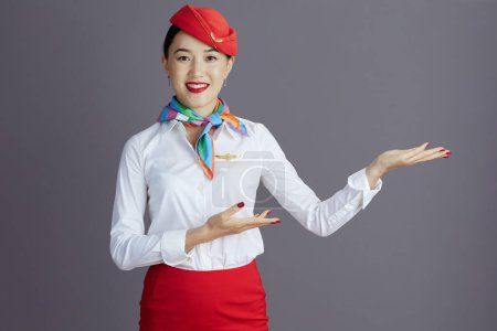 Photo for Smiling elegant air hostess asian woman in red skirt and hat uniform welcoming isolated on gray background. - Royalty Free Image