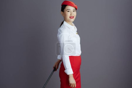 Foto de Feliz azafata moderna mujer asiática en falda roja y sombrero uniforme con bolsa de carro contra fondo gris. - Imagen libre de derechos