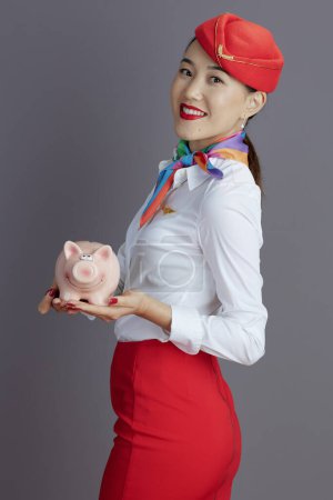 Photo for Smiling modern asian female stewardess in red skirt and hat uniform with piggy bank isolated on grey background. - Royalty Free Image