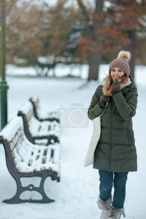 Foto de Sonriente mujer moderna de 40 años en abrigo verde y sombrero marrón al aire libre en el parque de la ciudad en invierno con manoplas y gorro. - Imagen libre de derechos