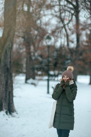 Téléchargez les photos : Femme moderne souriante en manteau vert et chapeau brun à l'extérieur dans le parc de la ville en hiver avec mitaines et bonnet. - en image libre de droit