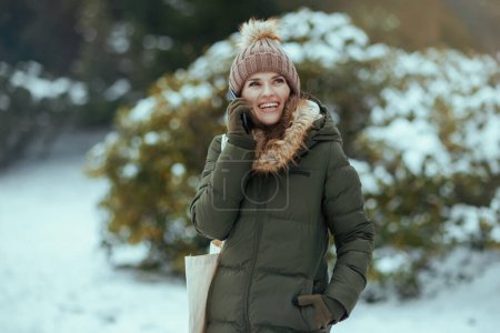 Téléchargez les photos : Joyeuse femme moderne en manteau vert et chapeau marron à l'extérieur dans le parc de la ville en hiver avec des mitaines et un bonnet près des branches enneigées parlant sur un smartphone. - en image libre de droit