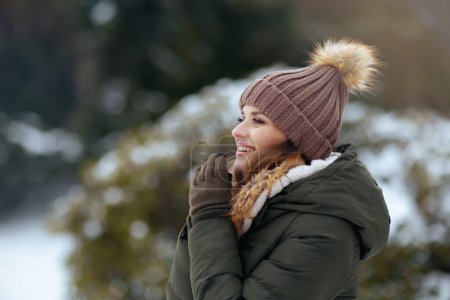 Photo for Smiling modern female in green coat and brown hat outdoors in the city park in winter with mittens and beanie hat. - Royalty Free Image
