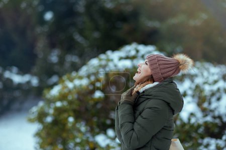 Foto de Sonriente mujer moderna de 40 años con abrigo verde y sombrero marrón al aire libre en el parque de la ciudad en invierno con manoplas y gorro cerca de ramas nevadas. - Imagen libre de derechos