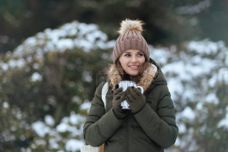 Photo for Happy modern woman in green coat and brown hat outdoors in the city park in winter with mittens, cup of soy latte and beanie hat near snowy branches. - Royalty Free Image