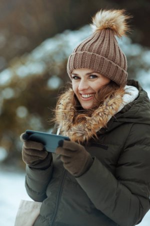 Téléchargez les photos : Heureuse femme moderne en manteau vert et chapeau brun à l'extérieur dans le parc de la ville en hiver avec des mitaines et bonnet chapeau envoyer un message texte en utilisant smartphone. - en image libre de droit