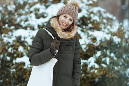 Foto de Feliz mujer moderna de 40 años en abrigo verde y sombrero marrón al aire libre en el parque de la ciudad en invierno con manoplas y gorro sombrero cerca de ramas nevadas. - Imagen libre de derechos