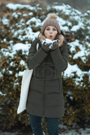 Foto de Mujer moderna en abrigo verde y sombrero marrón al aire libre en el parque de la ciudad en invierno con manoplas y gorro sombrero cerca de ramas nevadas. - Imagen libre de derechos