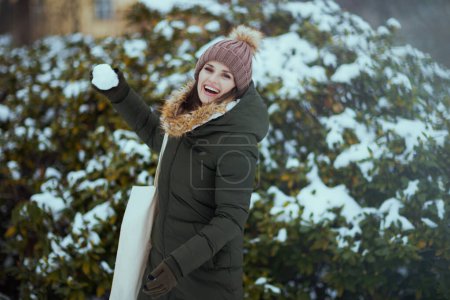 Foto de Mujer moderna feliz en abrigo verde y sombrero marrón al aire libre en el parque de la ciudad en invierno con manoplas y gorro de gorro lanzando bolas de nieve cerca de ramas nevadas. - Imagen libre de derechos