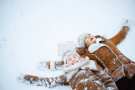 Photo for Smiling modern mother and daughter in coat, hat, scarf and mittens laying in snow outdoors in the city park in winter. - Royalty Free Image