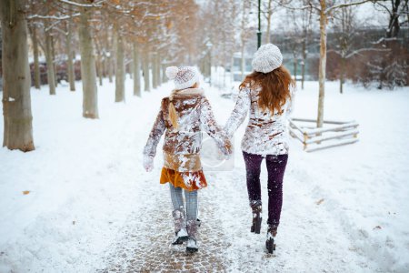 Téléchargez les photos : Vu de derrière mère et enfant modernes en manteau, chapeau, écharpe et mitaines en vêtements enneigés marchant à l'extérieur dans le parc de la ville en hiver. - en image libre de droit