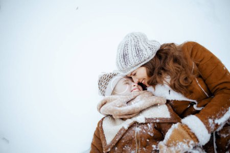 Photo for Happy elegant mother and daughter in coat, hat, scarf and mittens laying in snow outdoors in the city park in winter. - Royalty Free Image