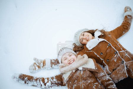 Téléchargez les photos : Mère et enfant élégants souriants en manteau, chapeau, écharpe et mitaines faisant ange de neige à l'extérieur dans le parc de la ville en hiver. - en image libre de droit