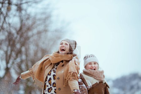 Téléchargez les photos : Heureuse élégante mère et fille en manteau, chapeau, écharpe et mitaines attraper la neige à l'extérieur en ville en hiver. - en image libre de droit