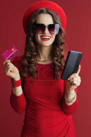 Téléchargez les photos : Joyeux Valentin. heureuse femme d'âge moyen moderne avec de longs cheveux ondulés en robe rouge et béret sur fond rouge avec carte de crédit et smartphone faire des achats en ligne sur le site e-commerce. - en image libre de droit