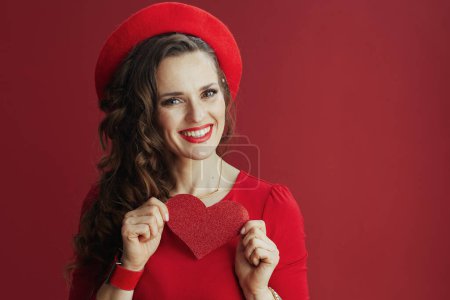Foto de Feliz San Valentín. Retrato de mujer de mediana edad elegante feliz en vestido rojo y boina sobre fondo rojo con corazón rojo y pelo largo ondulado. - Imagen libre de derechos