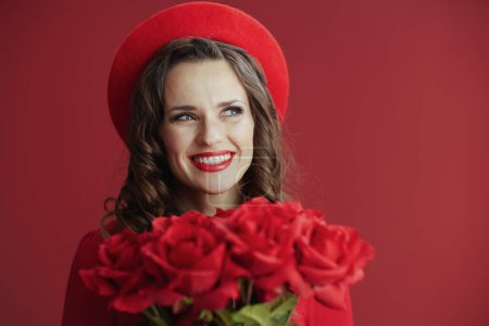 Foto de Feliz San Valentín. Retrato de mujer de 40 años con estilo sonriente en vestido rojo y boina sobre fondo rojo con rosas rojas. - Imagen libre de derechos
