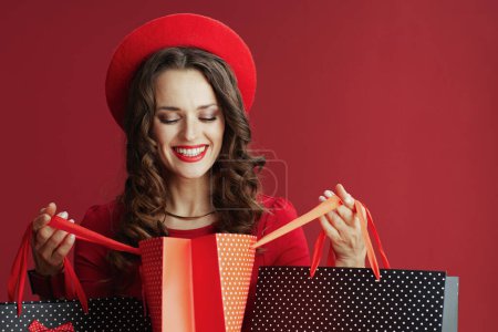 Foto de Feliz San Valentín. sonriente mujer moderna en vestido rojo y boina con bolsas de compras aisladas en rojo. - Imagen libre de derechos