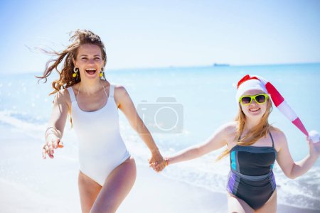 Foto de Feliz madre y el niño con estilo en sombrero de santa en la playa en traje de baño caminando. - Imagen libre de derechos
