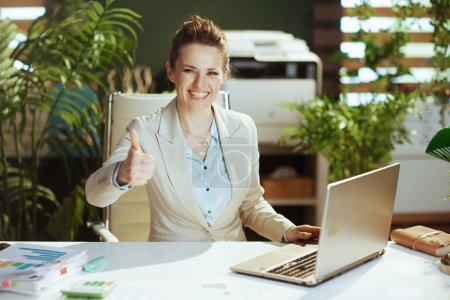 Photo for Portrait of happy modern woman worker in a light business suit in modern green office with laptop showing thumbs up. - Royalty Free Image