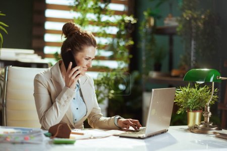Téléchargez les photos : Travailleuse moderne souriante dans un costume d'affaires léger dans un bureau vert moderne avec ordinateur portable parlant sur un smartphone. - en image libre de droit