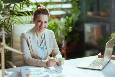 Foto de Retrato de mujer de negocios moderna sonriente en un traje de negocios ligero en la oficina verde moderna con computadora portátil. - Imagen libre de derechos