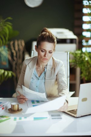 Foto de Moderna mujer de mediana edad trabajadora en un traje de negocios ligero en la oficina verde moderna con documentos. - Imagen libre de derechos
