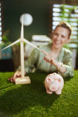 Photo for Eco Business. Closeup on smiling business woman in green blouse in green office with piggy bank and windmill - Royalty Free Image