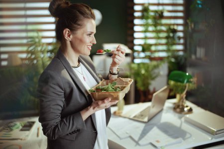 Téléchargez les photos : Un lieu de travail durable. heureuse femme d'âge moyen moderne employée dans un costume d'affaires gris dans un bureau vert moderne avec salade manger ordinateur portable. - en image libre de droit