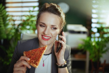 Téléchargez les photos : Un lieu de travail durable. souriant femme propriétaire de petite entreprise moderne dans un costume d'affaires gris dans un bureau vert moderne avec pizza parler sur un smartphone. - en image libre de droit