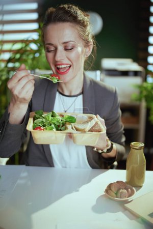 Photo for Sustainable workplace. happy modern middle aged small business owner woman in a grey business suit in modern green office eating salad. - Royalty Free Image